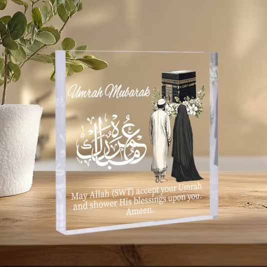 Decorative glass panel with religious text and images of a man and woman walking towards a Kaaba, on a wooden surface.
